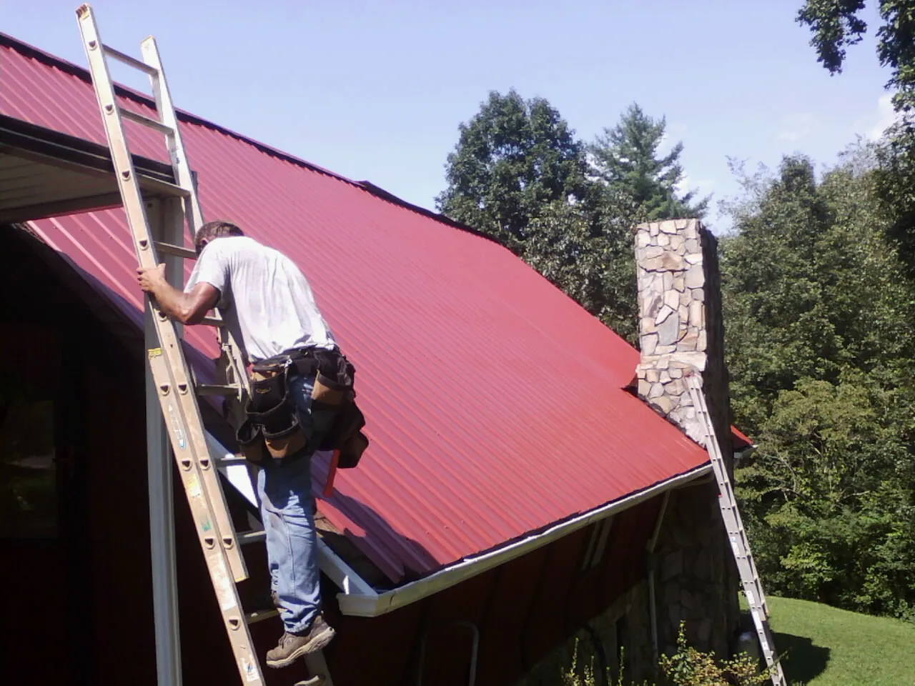Roofer on a ladder beside a red metal roof and stone chimney — Soffit & Fascia Repair by Roofing Brothers in Vacherie